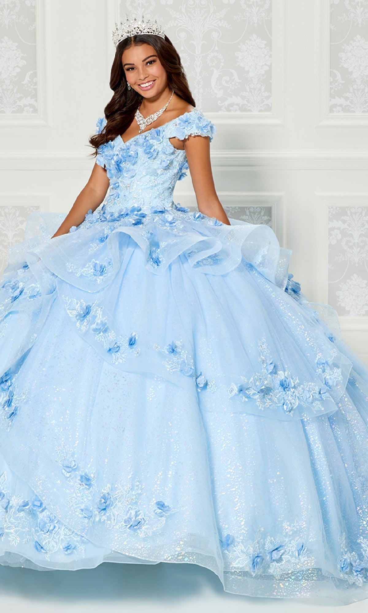 A young woman wearing the Princesa Quinceanera Dress PR30113, featuring blue off-the-shoulder floral appliqué, paired with a jeweled necklace and tiara, stands before a white patterned wall.